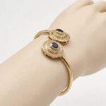 Natural oval blue sapphire and double diamond halo open cuff bangle bracelet in gold plated silver displayed on a model wrist